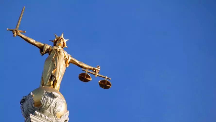 A picture of Lady Justice, a gold figure with arms outstretched holding a sword in one hand and scales in the other, against a clear blue sky