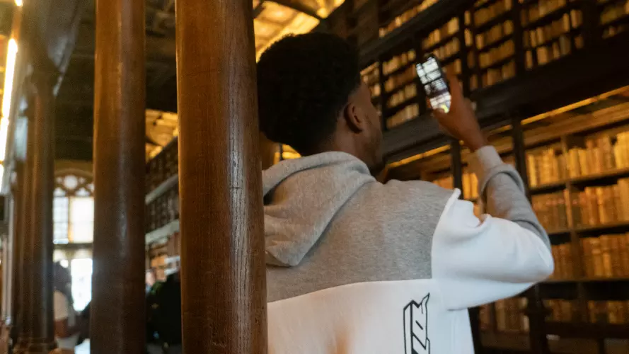 Person in a gray and white hoodie standing in a grand library with tall wooden columns and bookshelves. They are holding up a smartphone, possibly taking a photo or video. The library features an ornate ceiling, large windows letting in natural light, and