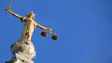 A picture of Lady Justice, a gold figure with arms outstretched holding a sword in one hand and scales in the other, against a clear blue sky