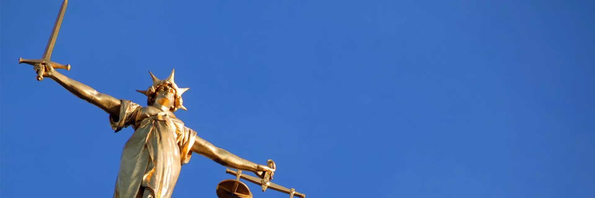 A picture of Lady Justice, a gold figure with arms outstretched holding a sword in one hand and scales in the other, against a clear blue sky